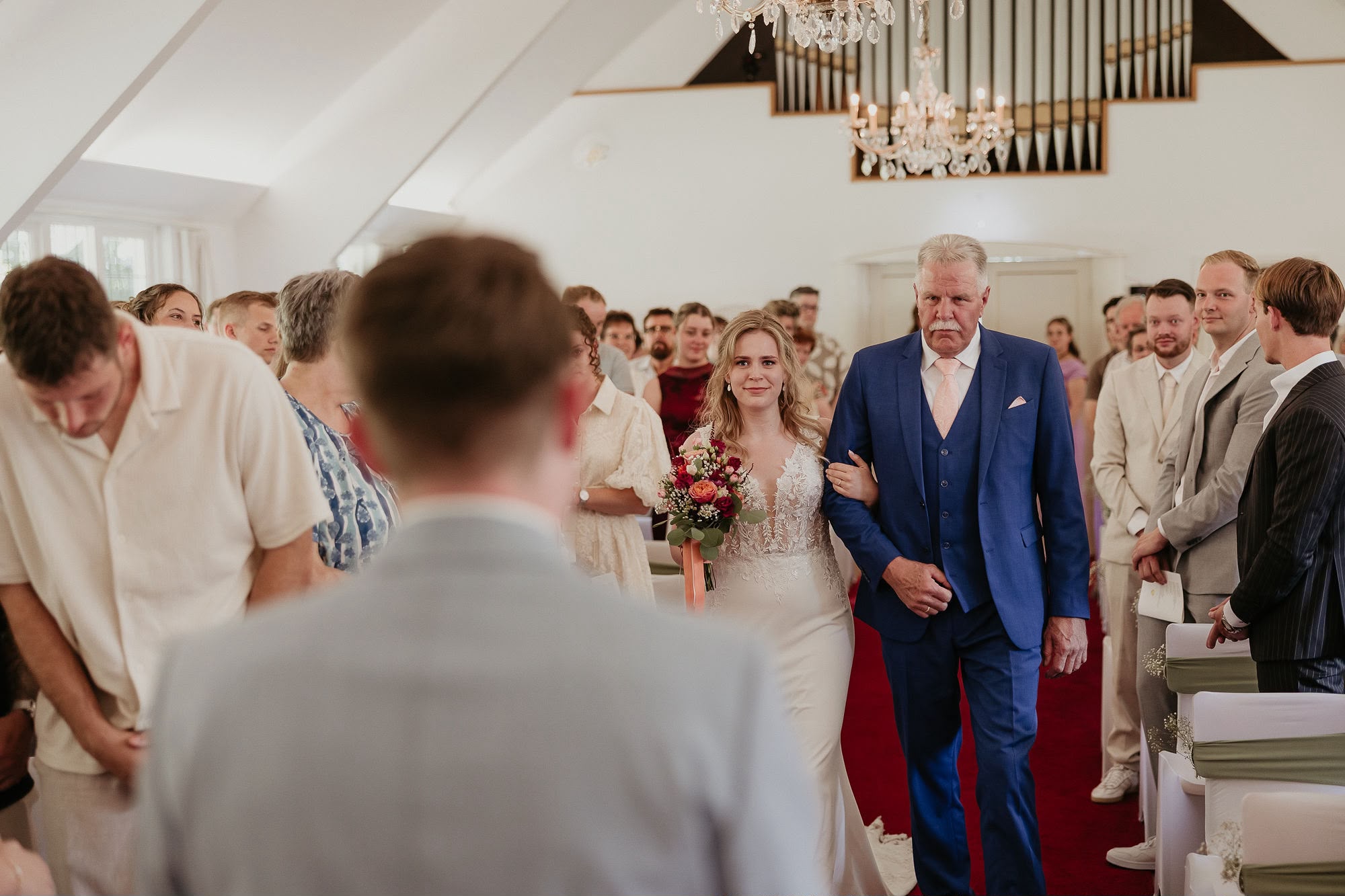 Bruid loopt gearmd met haar vader over de rode loper langs staande gasten