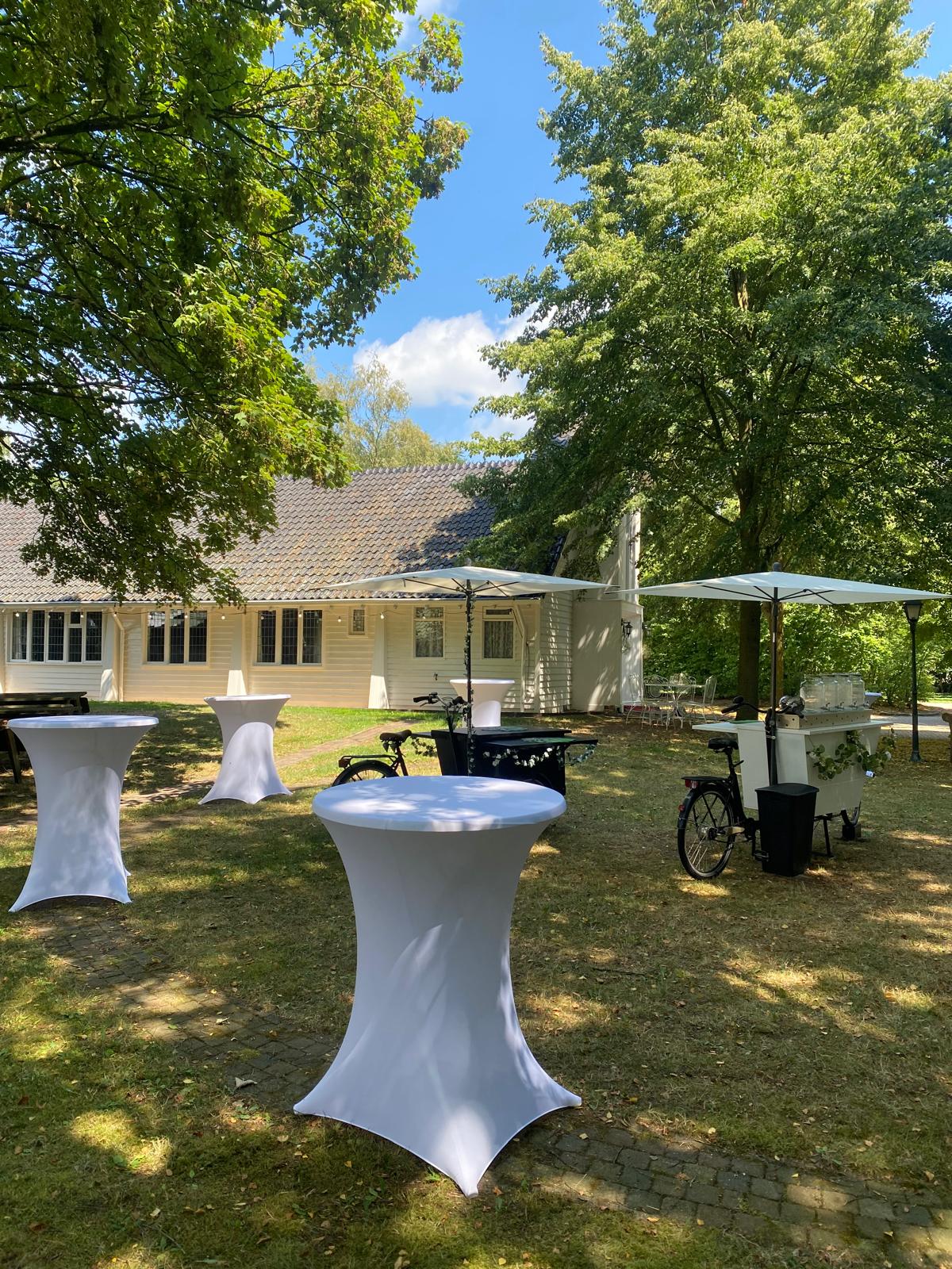 Witte statafels met parasols en een limonade bakfiets op het gazon naast het kerkje
