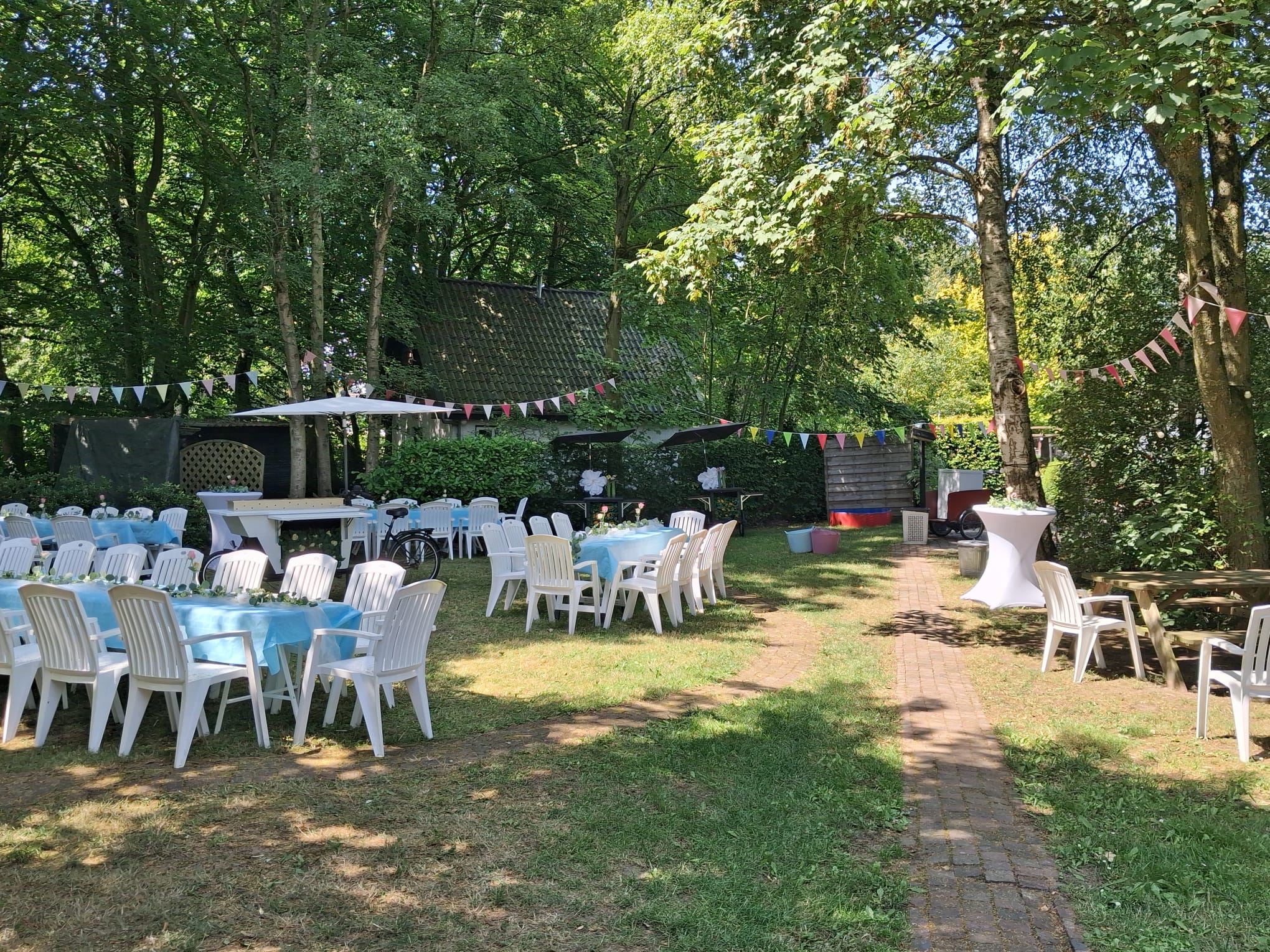 Tuinfeest bij Wit Kerkje met gedekte tafels, vlaggenlijnen en parasols onder oude bomen
